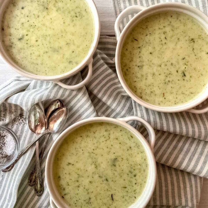 Broccoli Zucchini Soup in three white bowls with handles, on a striped dish towel, with three spoons and a small bowl of salt and pepper.