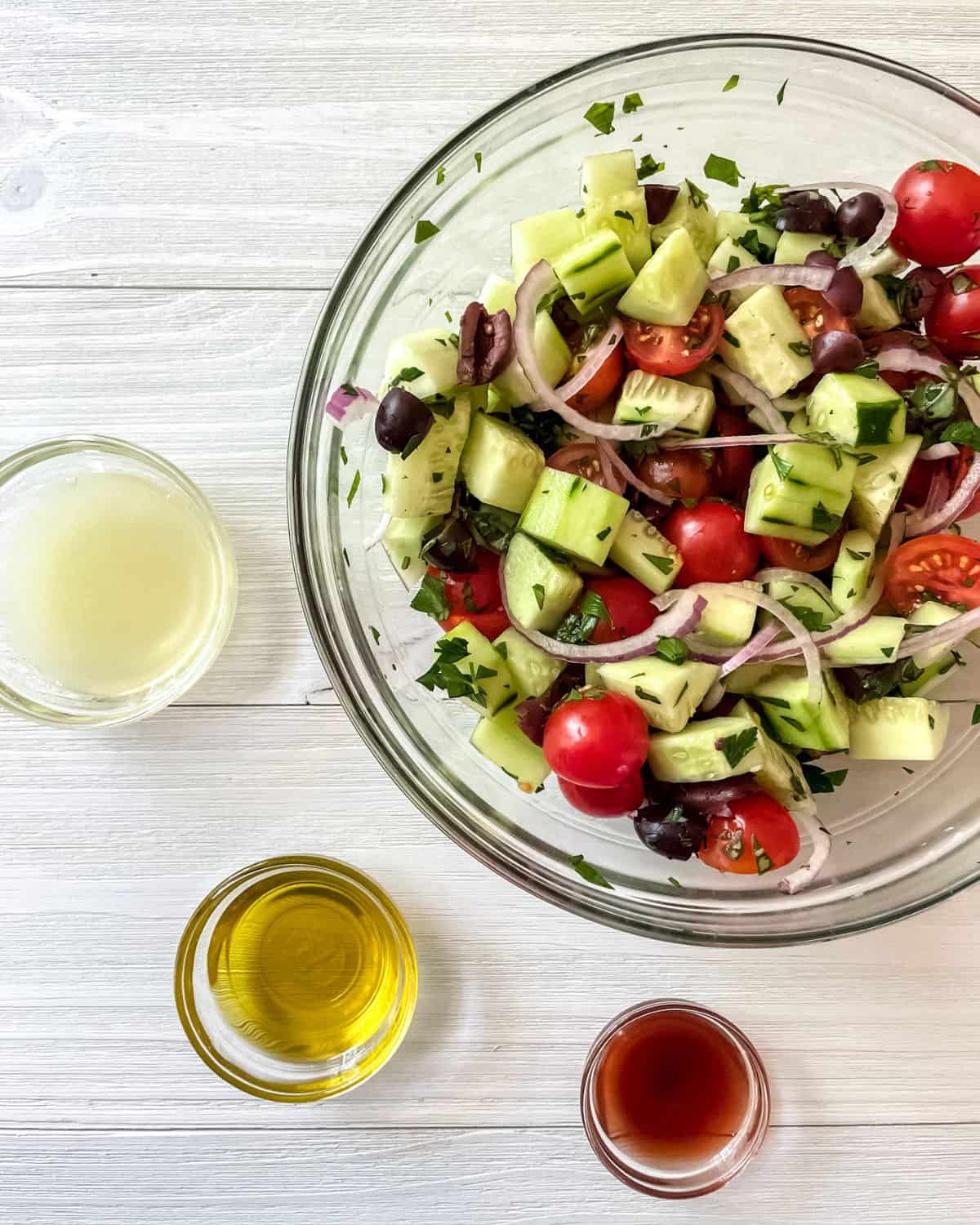 All dry tomato cucumber salad ingredients are in a glass bowl, with three small glass bowls next to it, containing fresh lemon juice, red wine vinegar and olive oil.