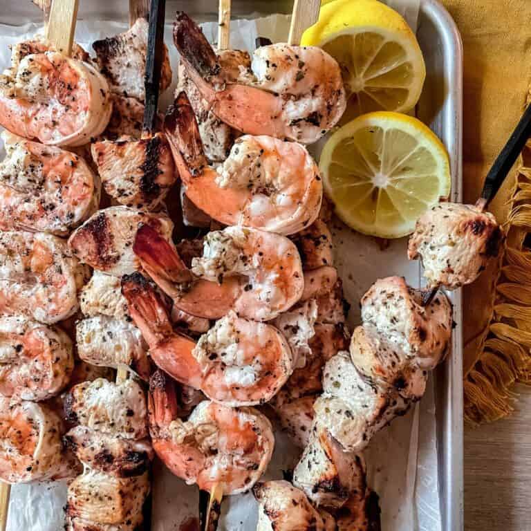 Grilled shrimp and chicken kabobs on a parchment lined sheet pan, lemon wedges in the top right corner and a yellow napkin in the top right corner.