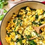 Zucchini, eggs, spinach and feta cheese scrambled in a red skillet with handles, scooped with a wooden spoon.