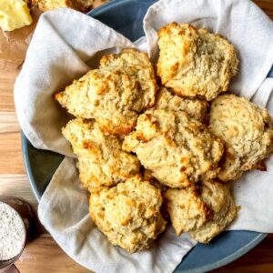 3 ingredient drop biscuits in a blue bowl lined with a white, cloth napkin, a measuring cup of flour is in the bottom left corner, butter and a crumbled biscuit is in the top left corner.
