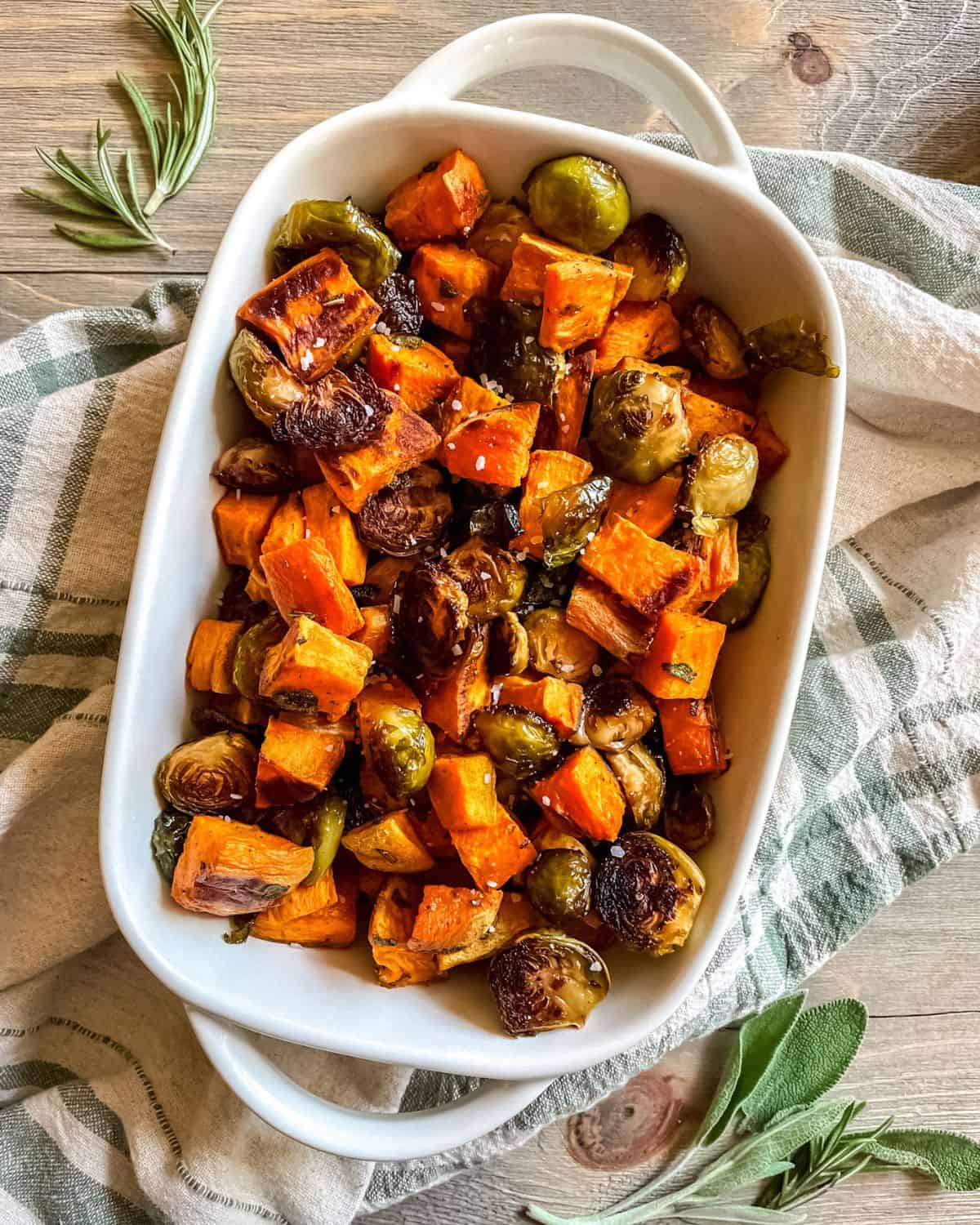 Roasted sweet potatoes and roasted brussel sprouts in glass casserole dish with handles, on a green and white dish towel, with fresh herbs on the sides.