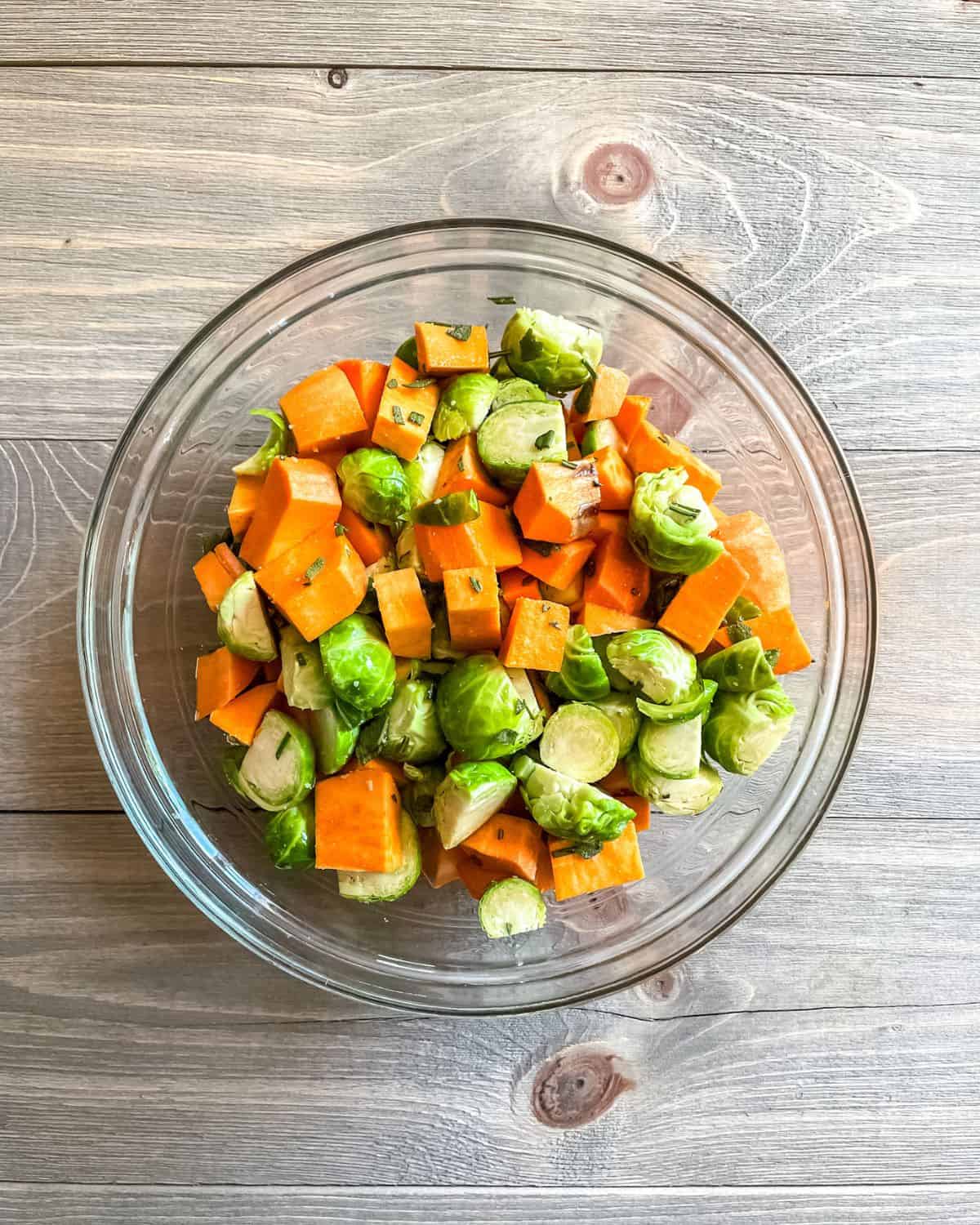 Sweet potatoes, brussel sprouts, fresh herbs and olive oil mixed in a glass bowl.