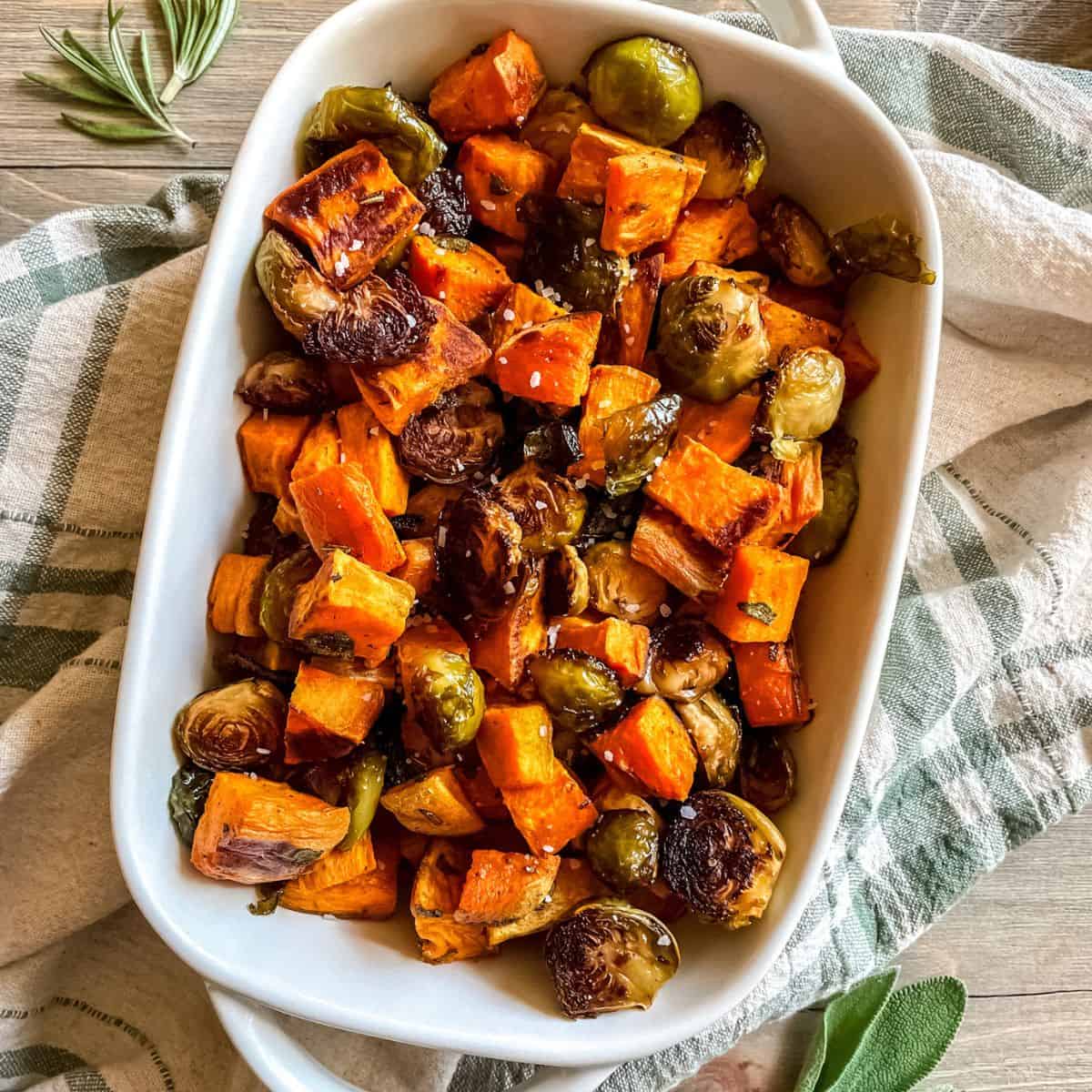 Roasted sweet potatoes and roasted brussel sprouts in glass casserole dish with handles, on a green and white dish towel, with fresh herbs on the sides.