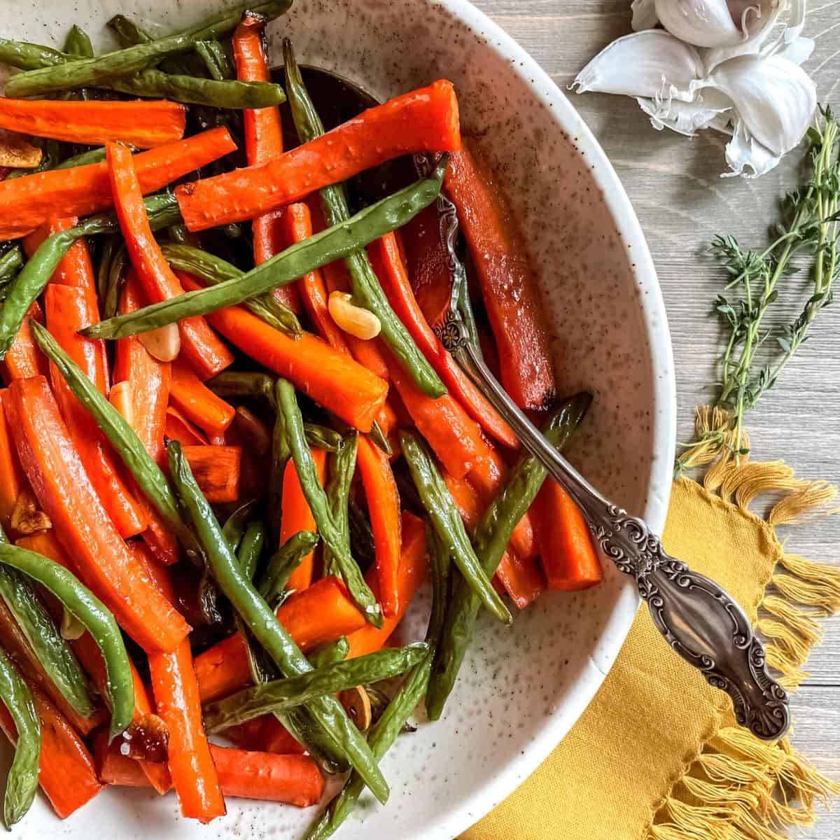 Roasted green beans and carrots in a round bowl, with a spoon, on a yellow cloth napkin, with garlic and fresh thyme in the background.