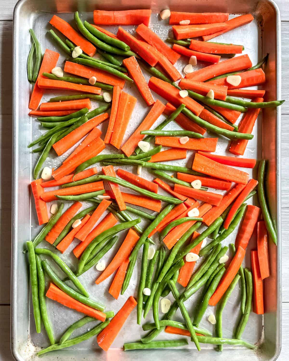 Sliced carrots and green beans on a large sheet pan with slices of garlic, uncooked.