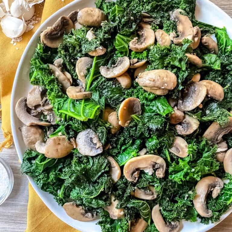 Sauteed Kale and Mushrooms on a white plate, sitting on top of a golden yellow napkin, with raw garlic cloves in the top left corner.