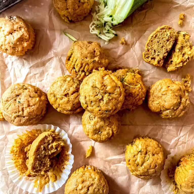Zucchini and pumpkin muffins on parchment paper, with a cheese grater in the top left corner and a partially grated zucchini in the top right corner.
