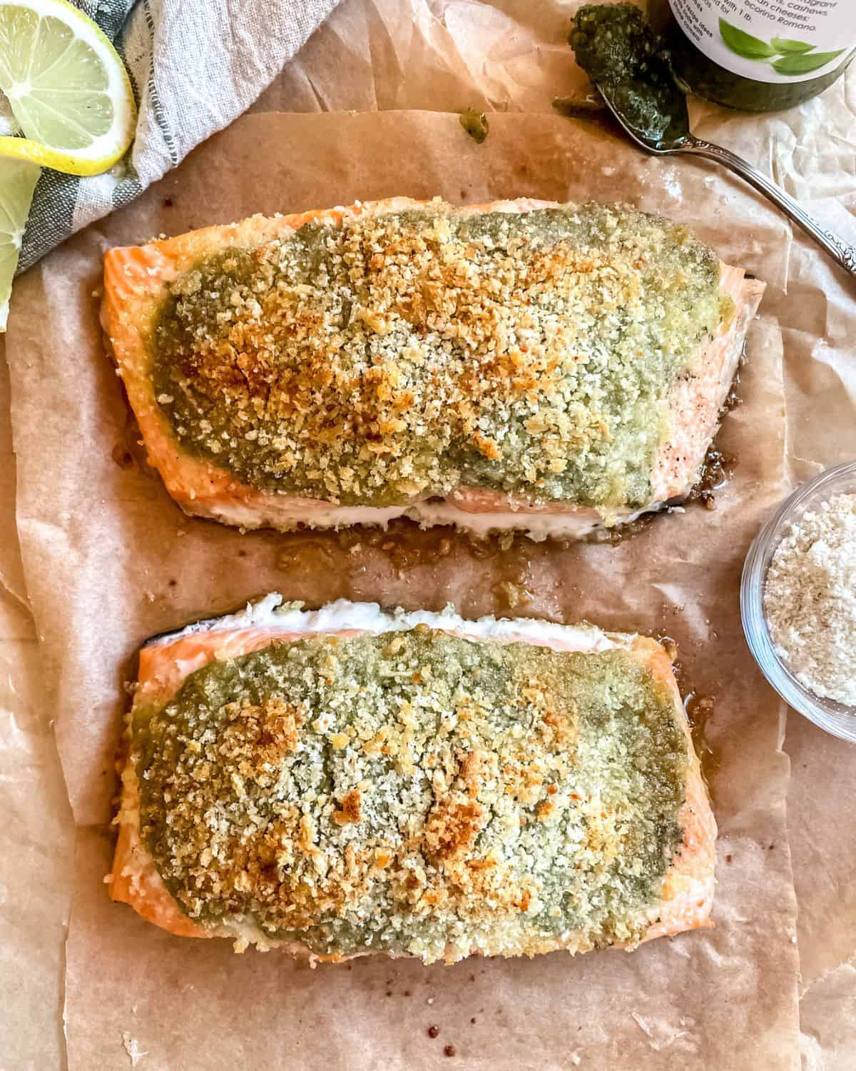 Pesto crusted salmon on layers of parchment paper, a jar of basil pesto and a spoon is in the top right corner, a dish of parmesan cheese is next to the salmon with a lemon wedge in the top left corner.