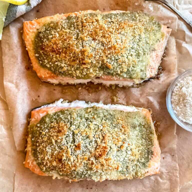 Pesto crusted salmon on layers of parchment paper, a dish of parmesan cheese is next to the salmon with a lemon wedge in the top left corner.