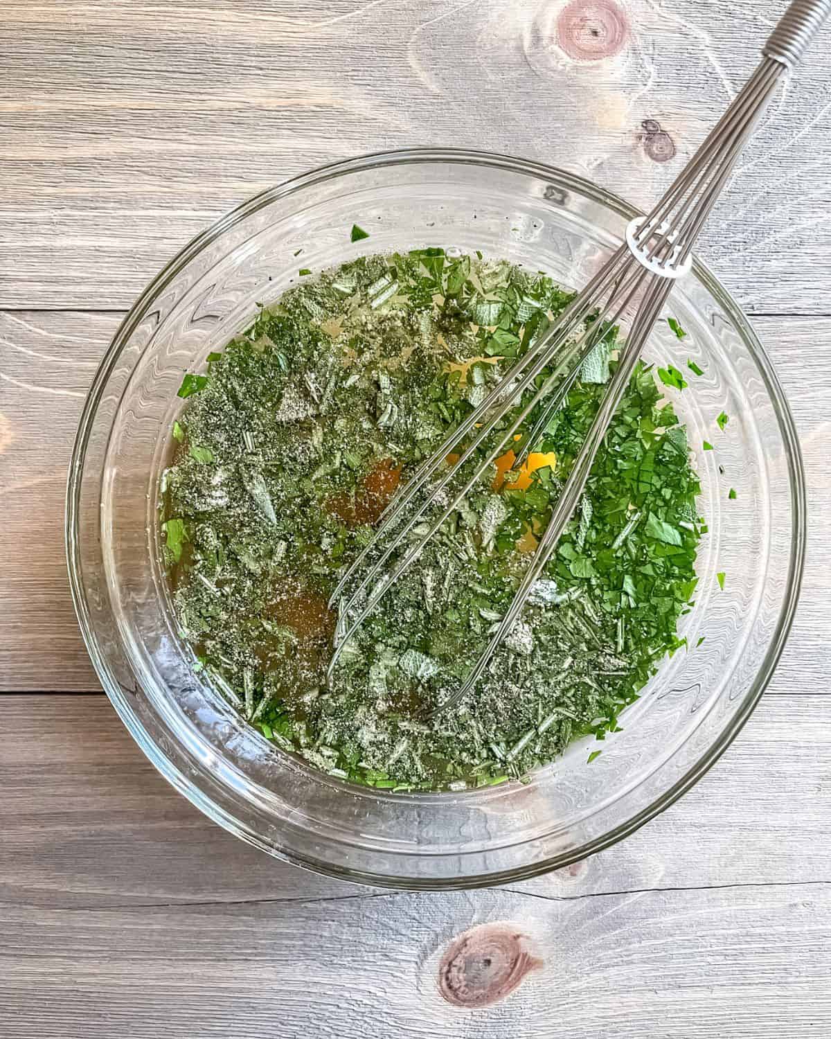 Liquid ingredients and chopped fresh herbs in a glass bowl with a whisk