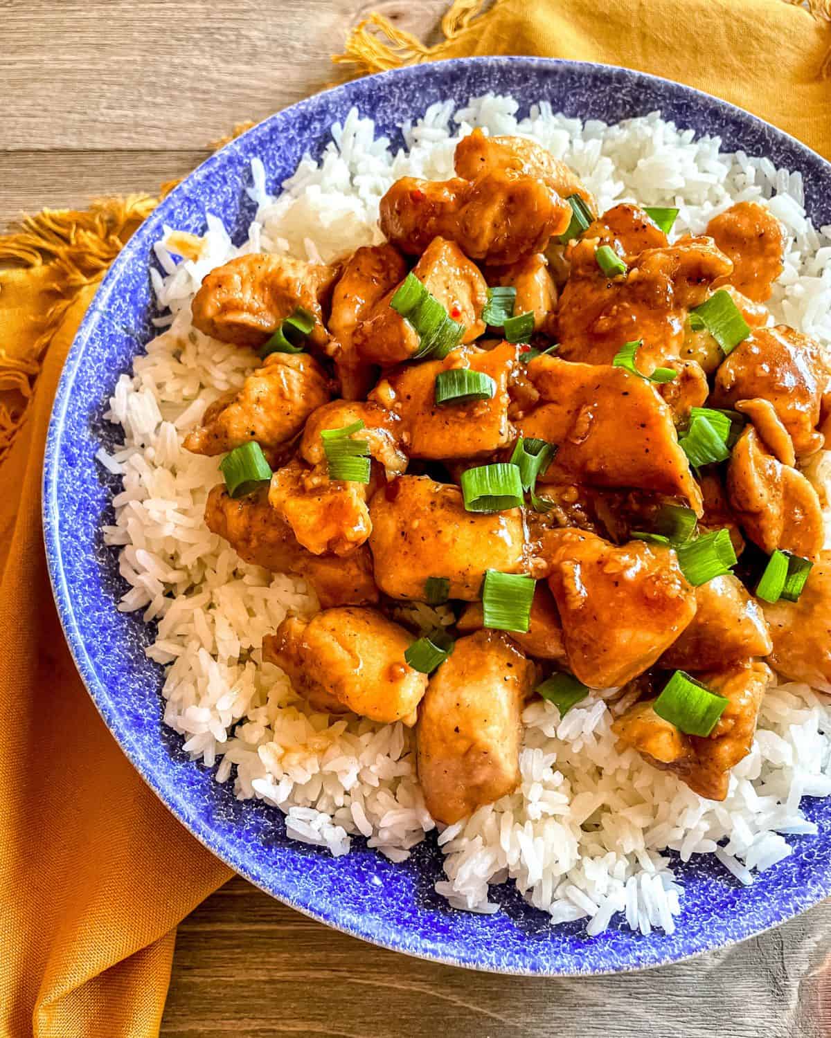 Honey Butter Garlic Chicken on a bed of white rice, in a blue bowl, garnished with green onion, on a yellow napkin.