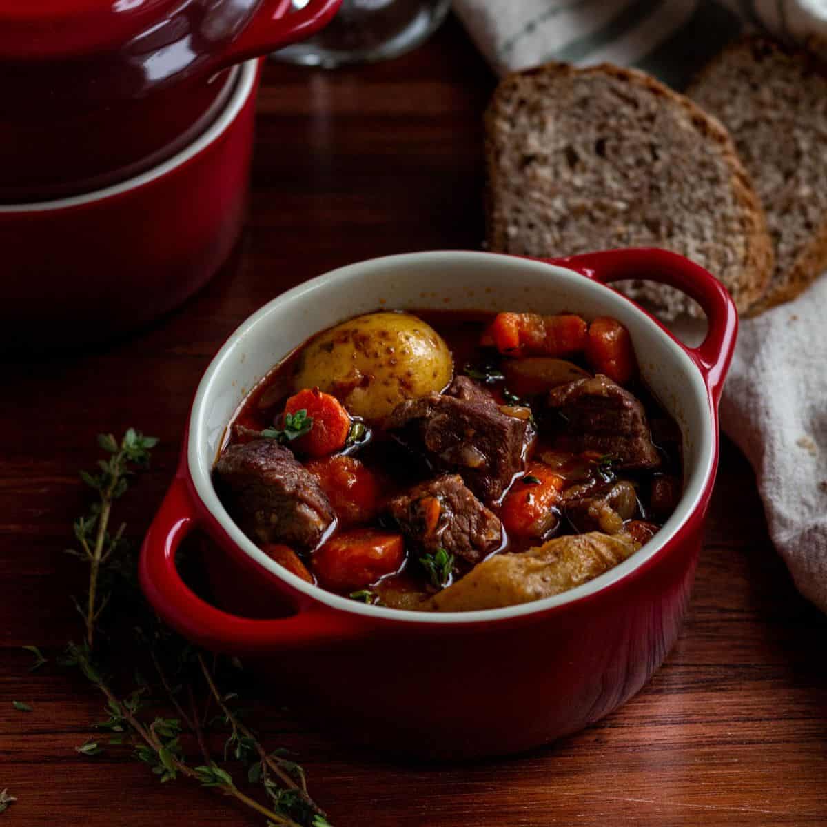 Red bowl of homemade beef stew. Bowl has handles. Fresh thyme and slices of bread surround the bowl.