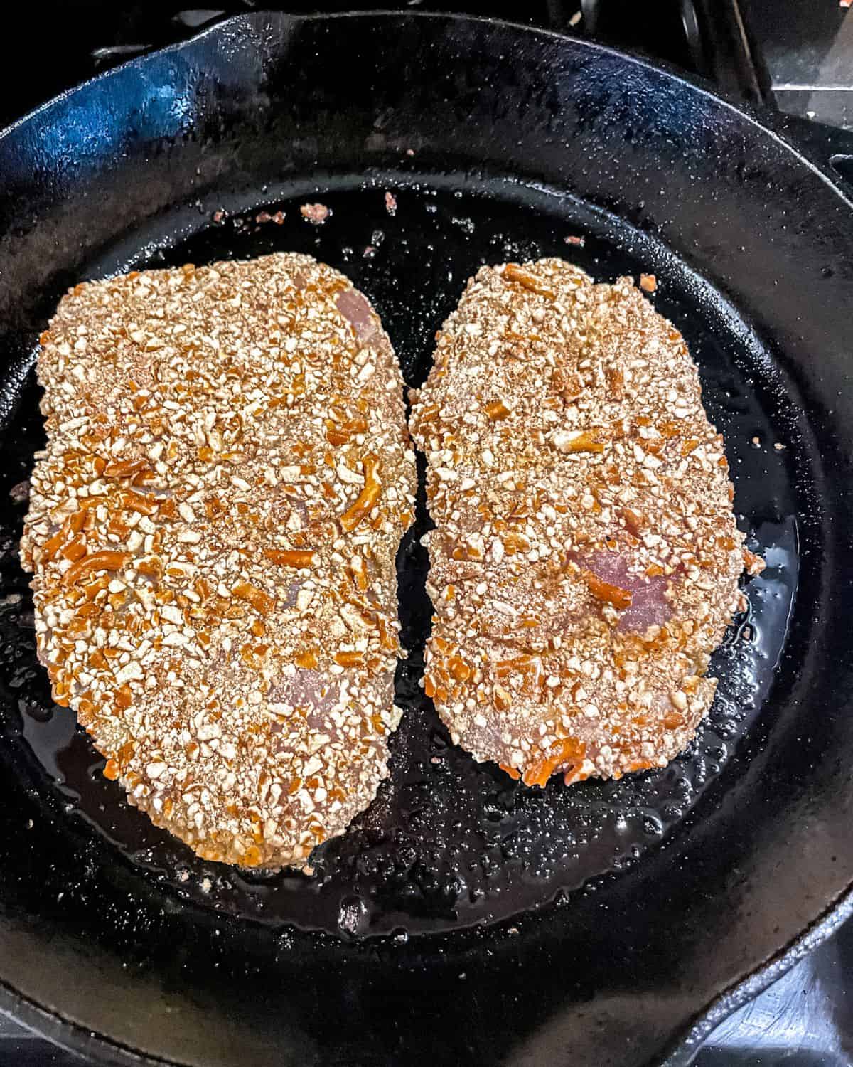 Pretzel chicken cutlets searing in a cast iron skillet.
