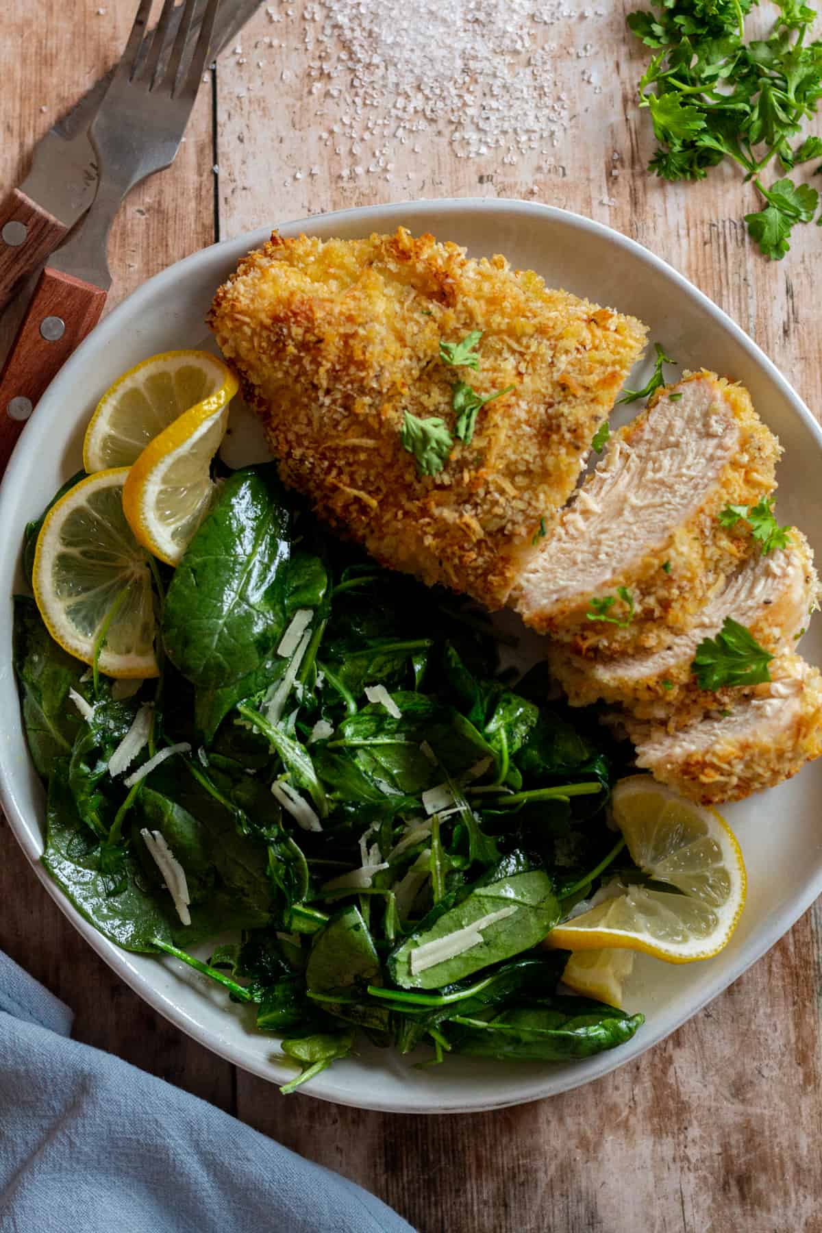 Baked Panko Chicken on a plate with sauteed greens and lemon slices, with a blue napkin and a fork and a knife.