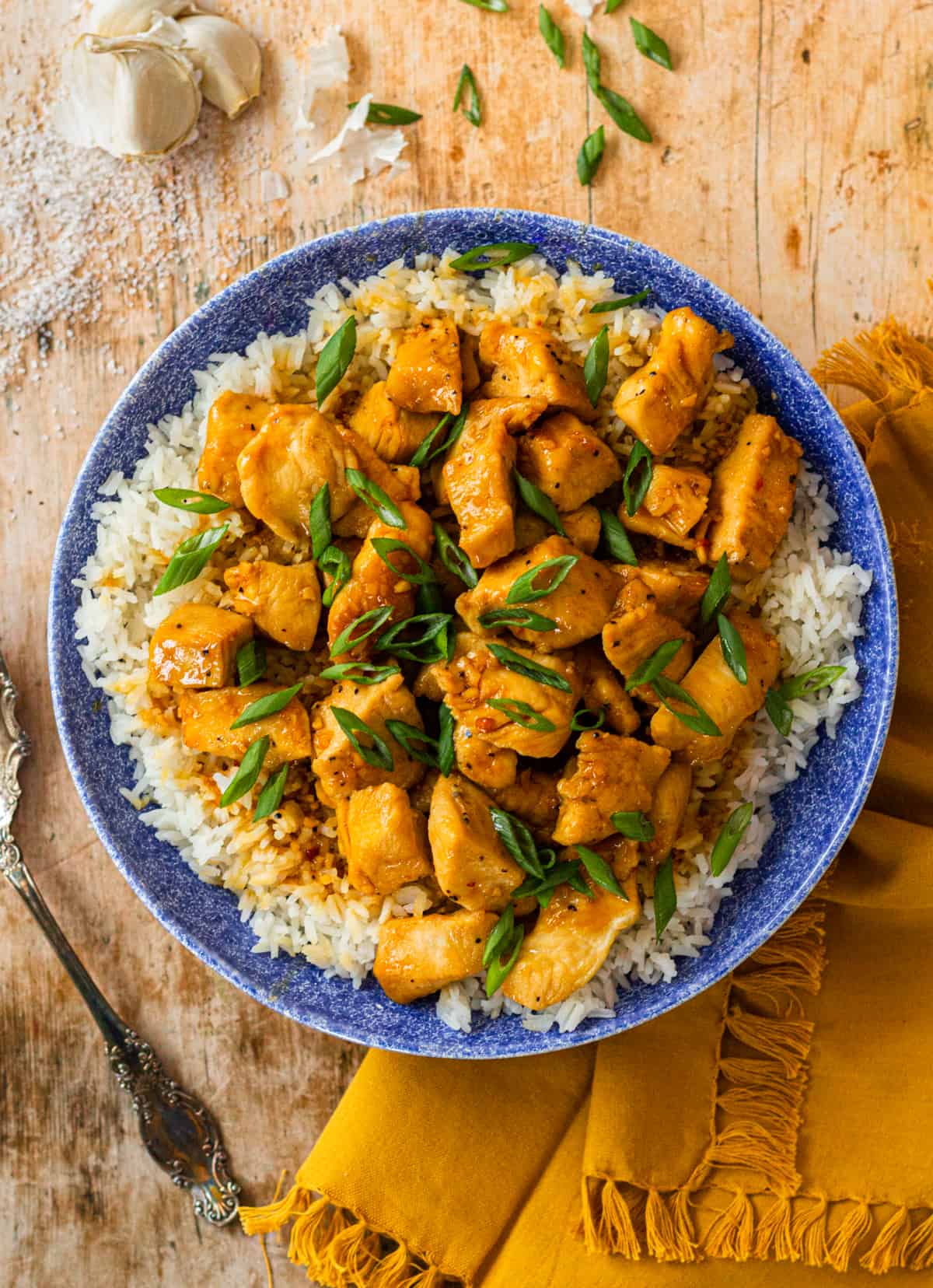 Honey Butter Garlic Chicken in a blue bowl, over a bed of white rice, on a yellow napkin with garlic, green onion and salt in the background.