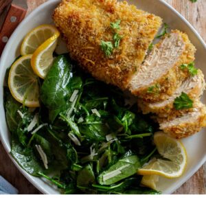 Baked panko chicken on a plate with greens and lemon wedges.