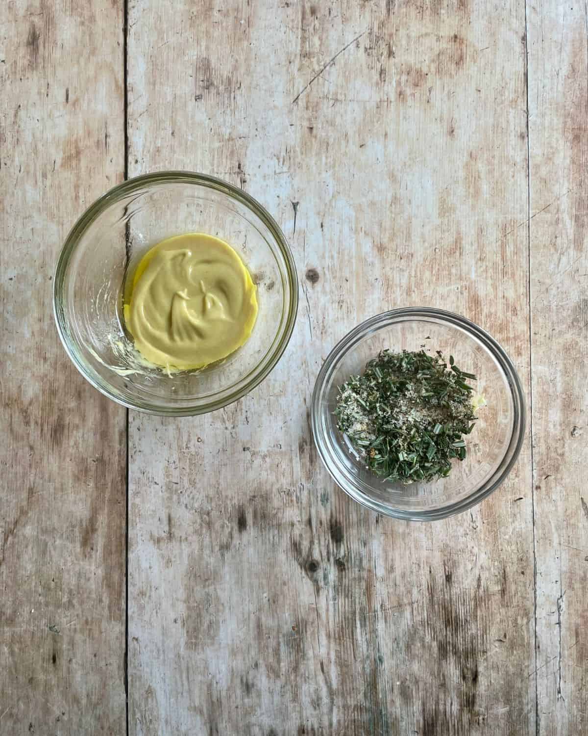 Small glass bowl of dijon mustard and olive oil, and a small glass bowl of dried and fresh herbs.