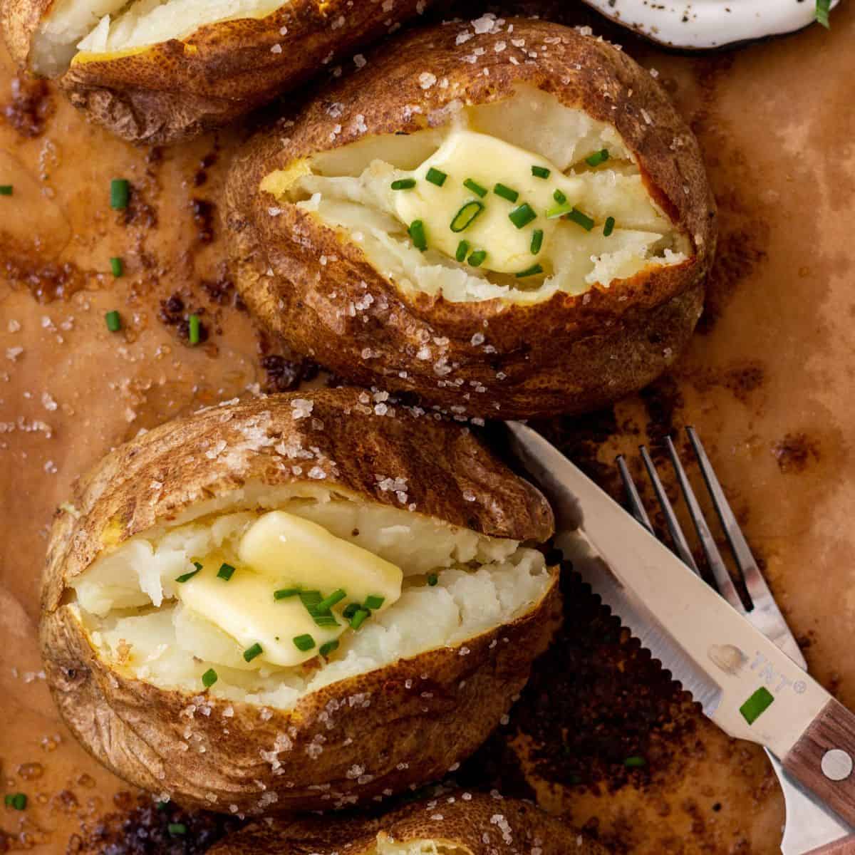 Two baked potatoes, cut open with butter and chives, on parchment paper with a fork and knife.