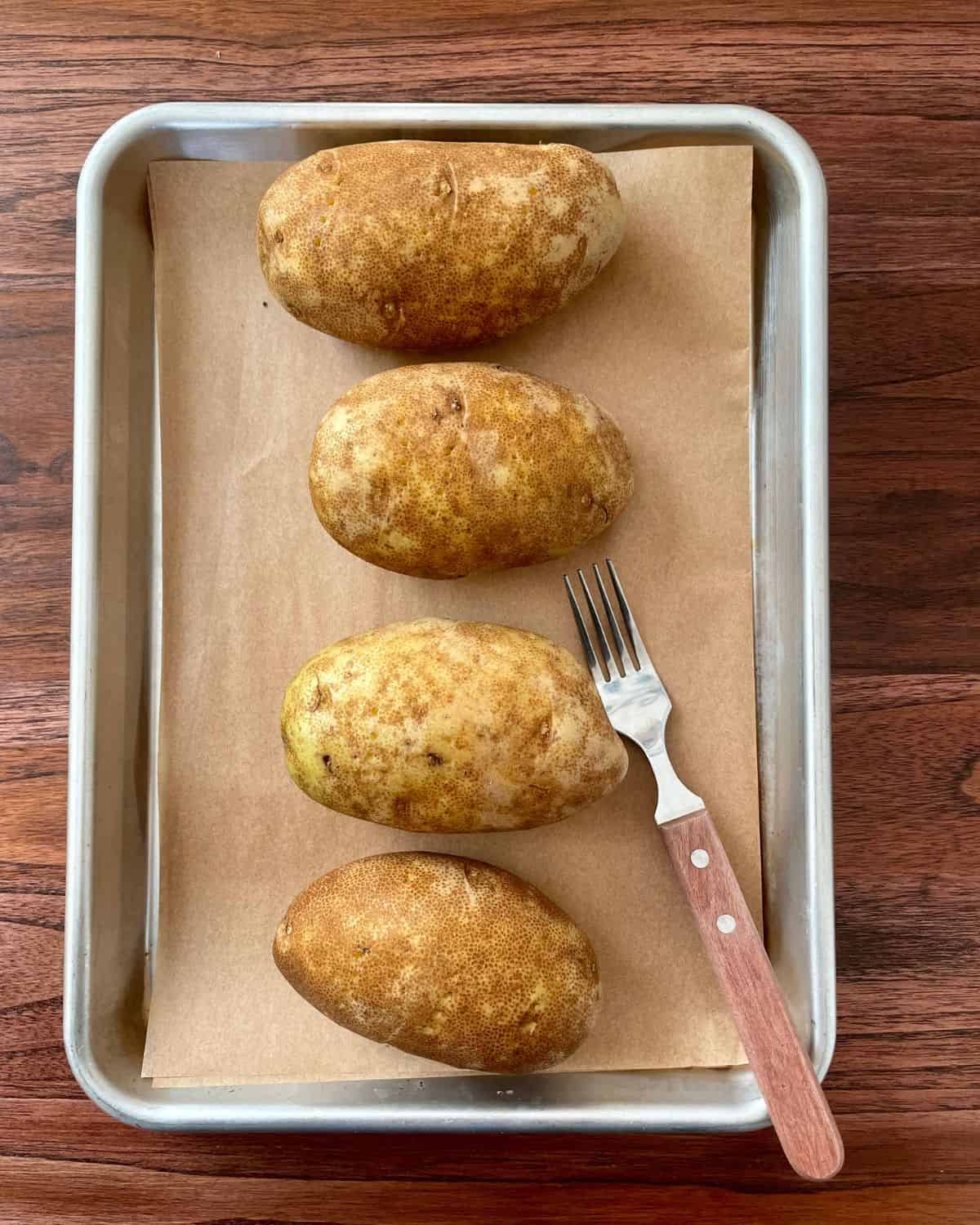 Four raw potatoes on a parchment lined baking sheet with a fork.