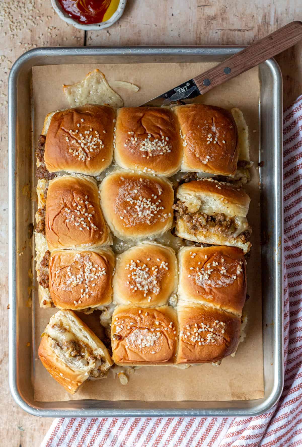 Cheeseburger Sliders with Hawaiian Rolls on a sheet pan lined with parchment paper, with a knife and a bowl of ketchup and mustard.