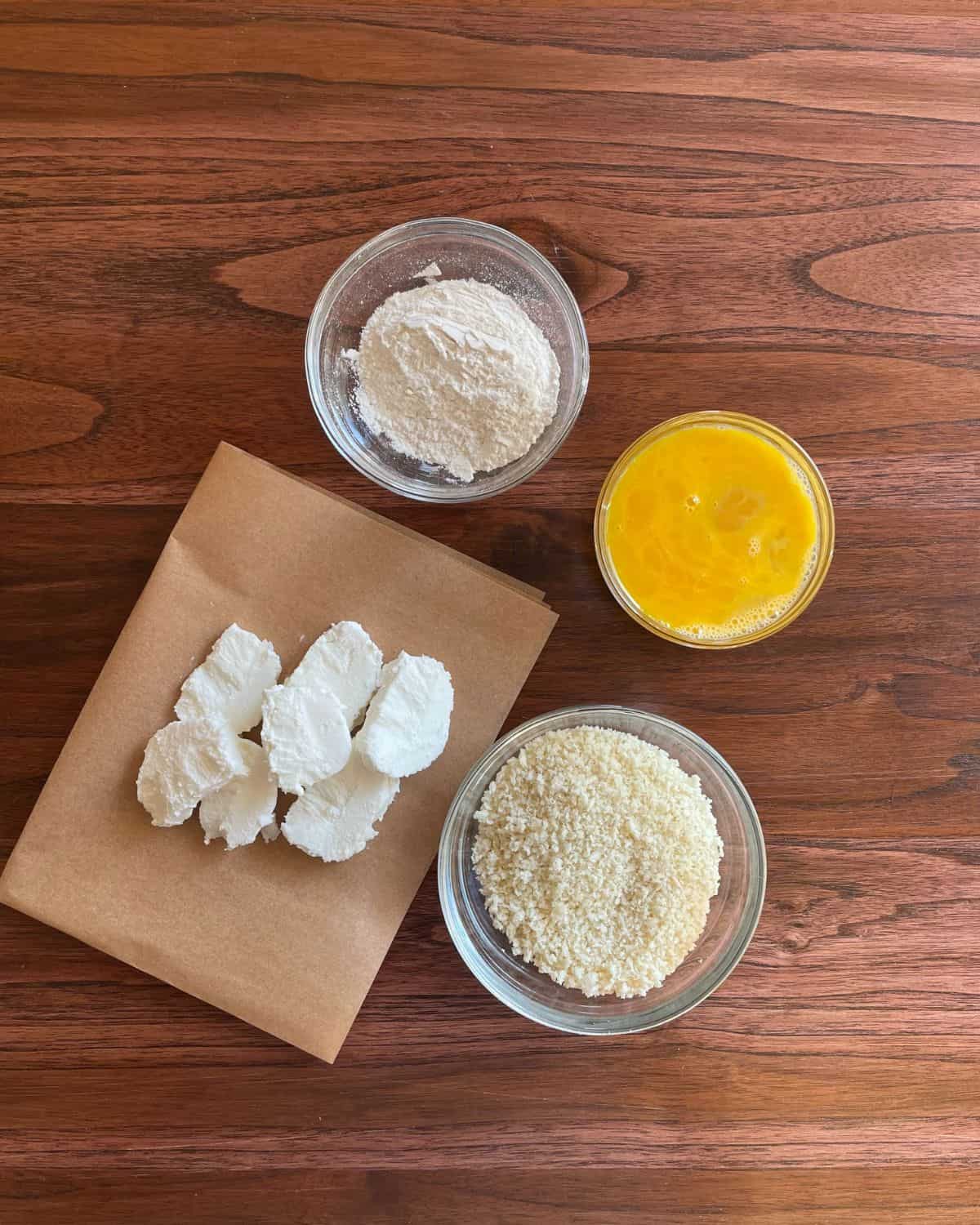 Sliced goat cheese rounds, a bowl of flour, a bowl of panko bread crumbs, and a bowl of whisked eggs.