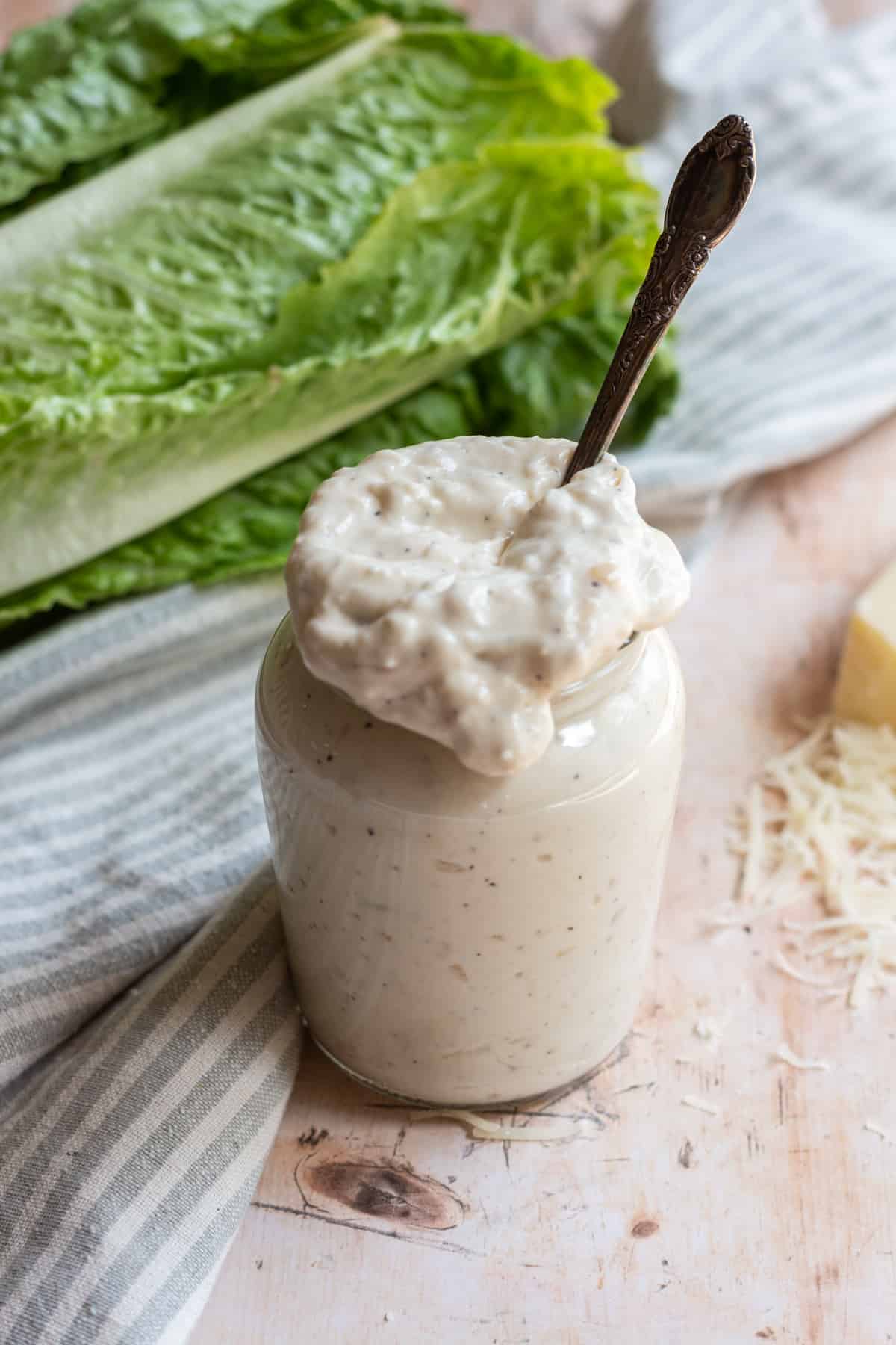 Small glass jar of Caesar dressing (no anchovies) with a spoon, lettuce leaves in the background, a striped towel in the background, and shredded parmesan cheese around the jar.