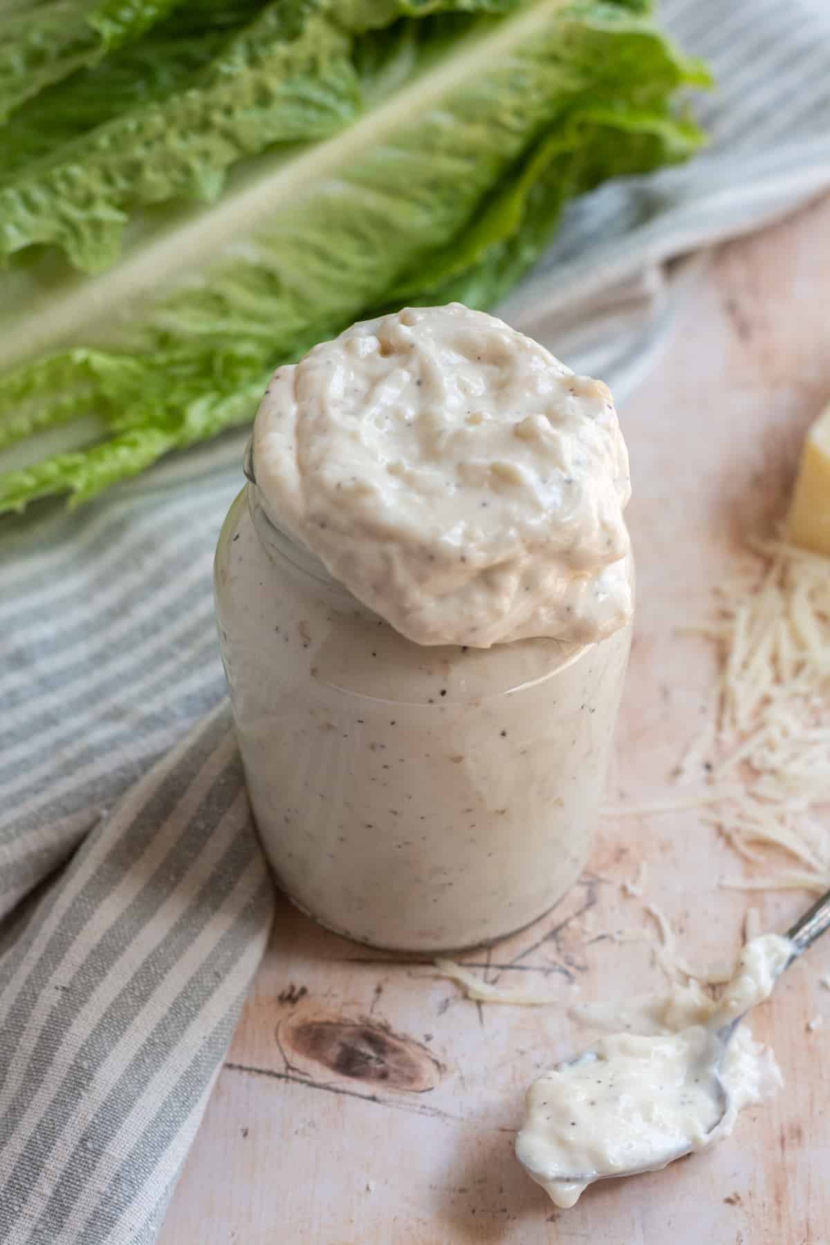 Small glass jar of Caesar dressing without anchovies with a spoon on the table, lettuce leaves in the background, a striped towel in the background, and shredded parmesan cheese around the jar.
