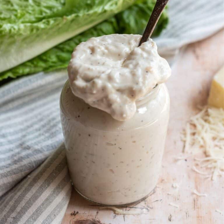 Small glass jar of Caesar dressing without anchovies with a spoon, lettuce leaves in the background, a striped towel in the background, and shredded parmesan cheese around the jar.