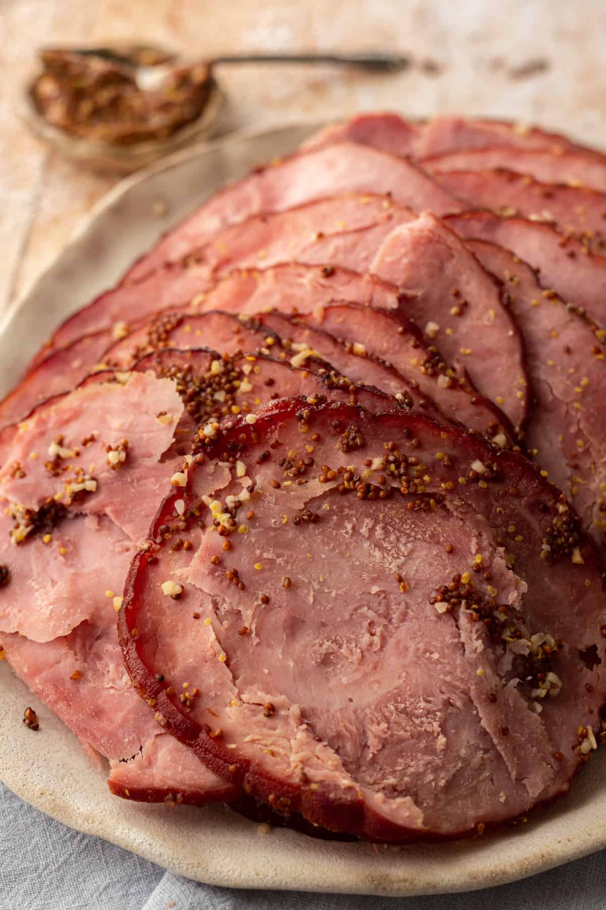 Up close image of sliced spiral ham on a beige, rustic style plate with dijon mustard in the background.