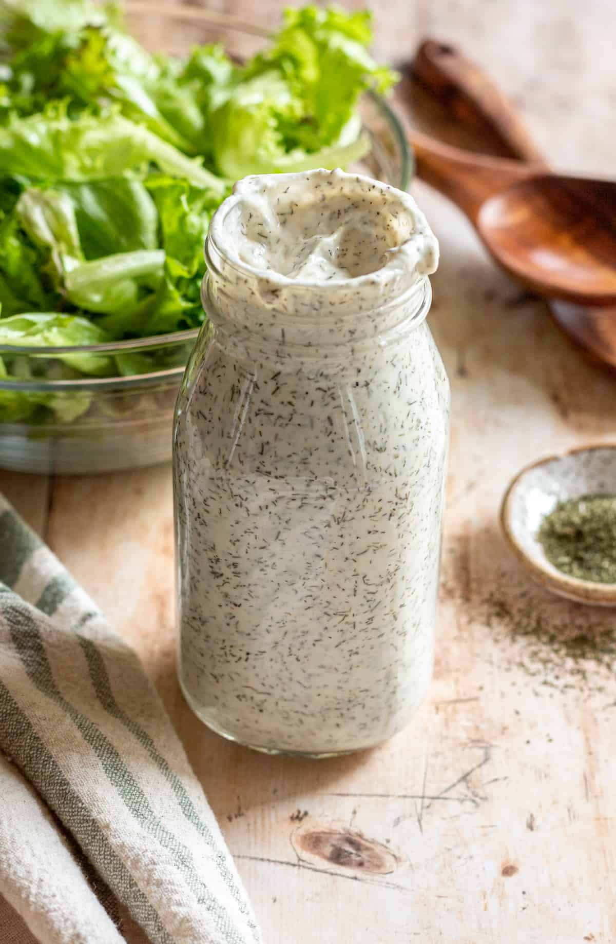 Creamy dill salad dressing in a glass jar, a bowl of lettuce and wooden salad spoons are in the background.