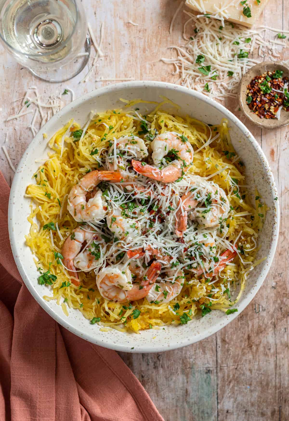 Grilled spaghetti squash shrimp scampi topped with parmesan cheese and red pepper flakes, in a bowl, with a pink napkin and a glass of white wine.