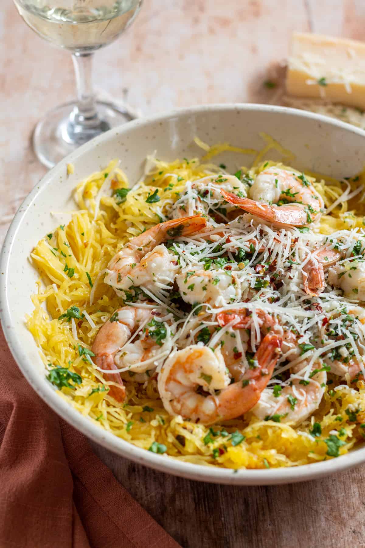 Grilled spaghetti squash shrimp scampi topped with parmesan cheese and red pepper flakes, in a bowl with a glass of white wine in the background.