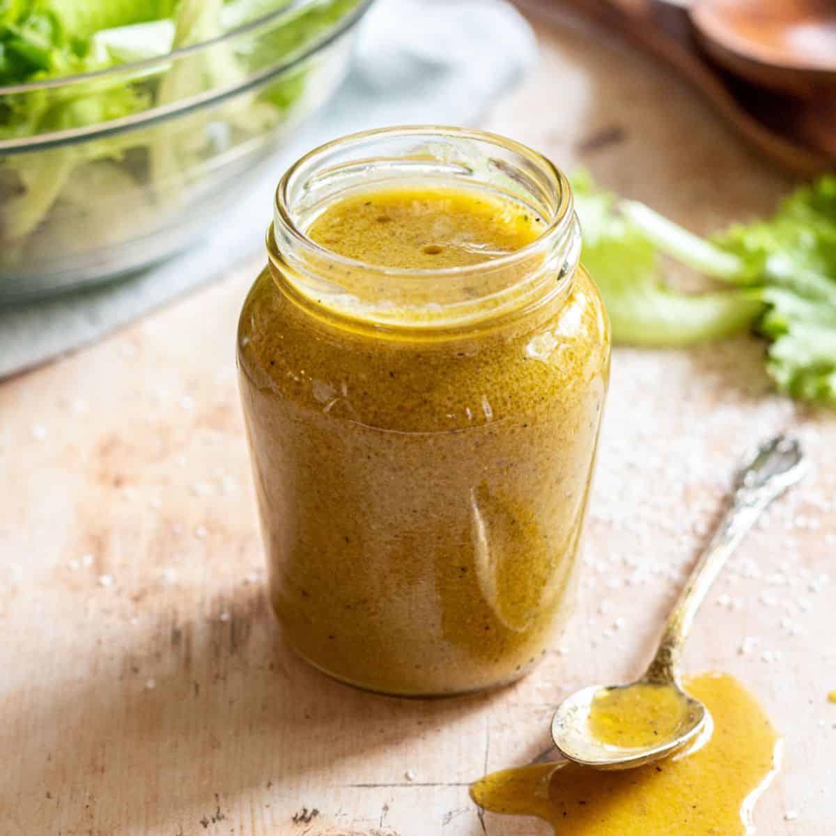 Maple Dijon Vinaigrette in a small glass jar with a spoon and some spilled dressing on the side, salad leaves in the background.