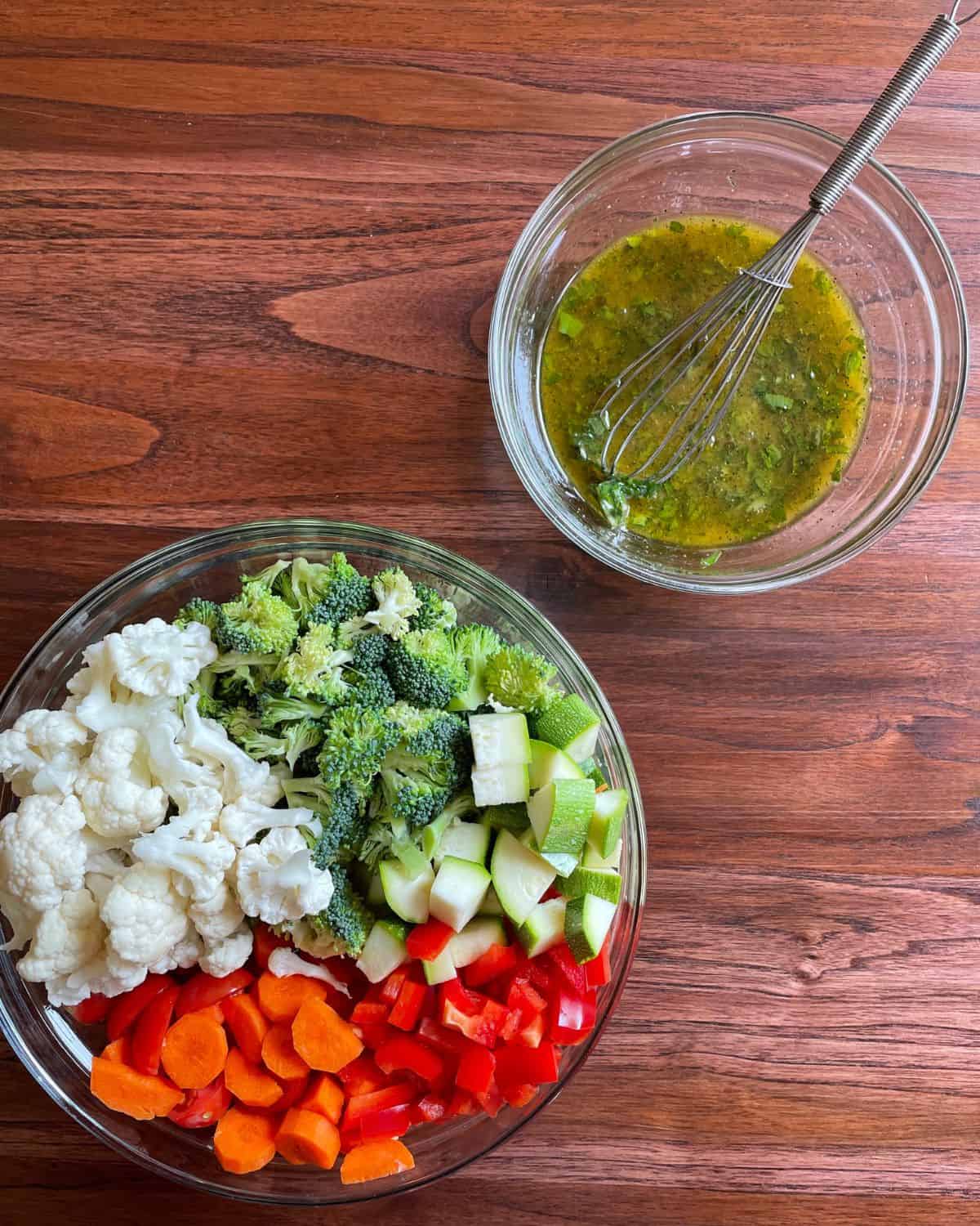 A large glass bowl of vegetables with the small glass bowl of salad dressing next to it.