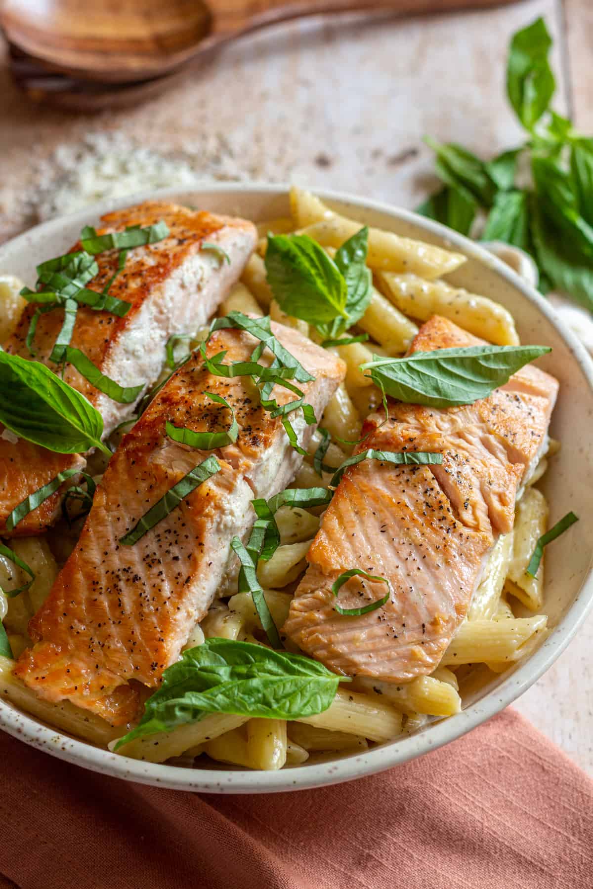 Three seared salmon filets over a bowl of penne-al-salmone pasta, garnished with fresh basil leaves. Fresh basil, wooden spoons, and parmesan cheese can be seen in the background.