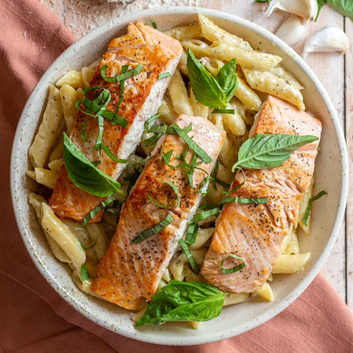 Three seared salmon filets over a bowl of Penne al Salmone pasta, garnished with fresh basil leaves.