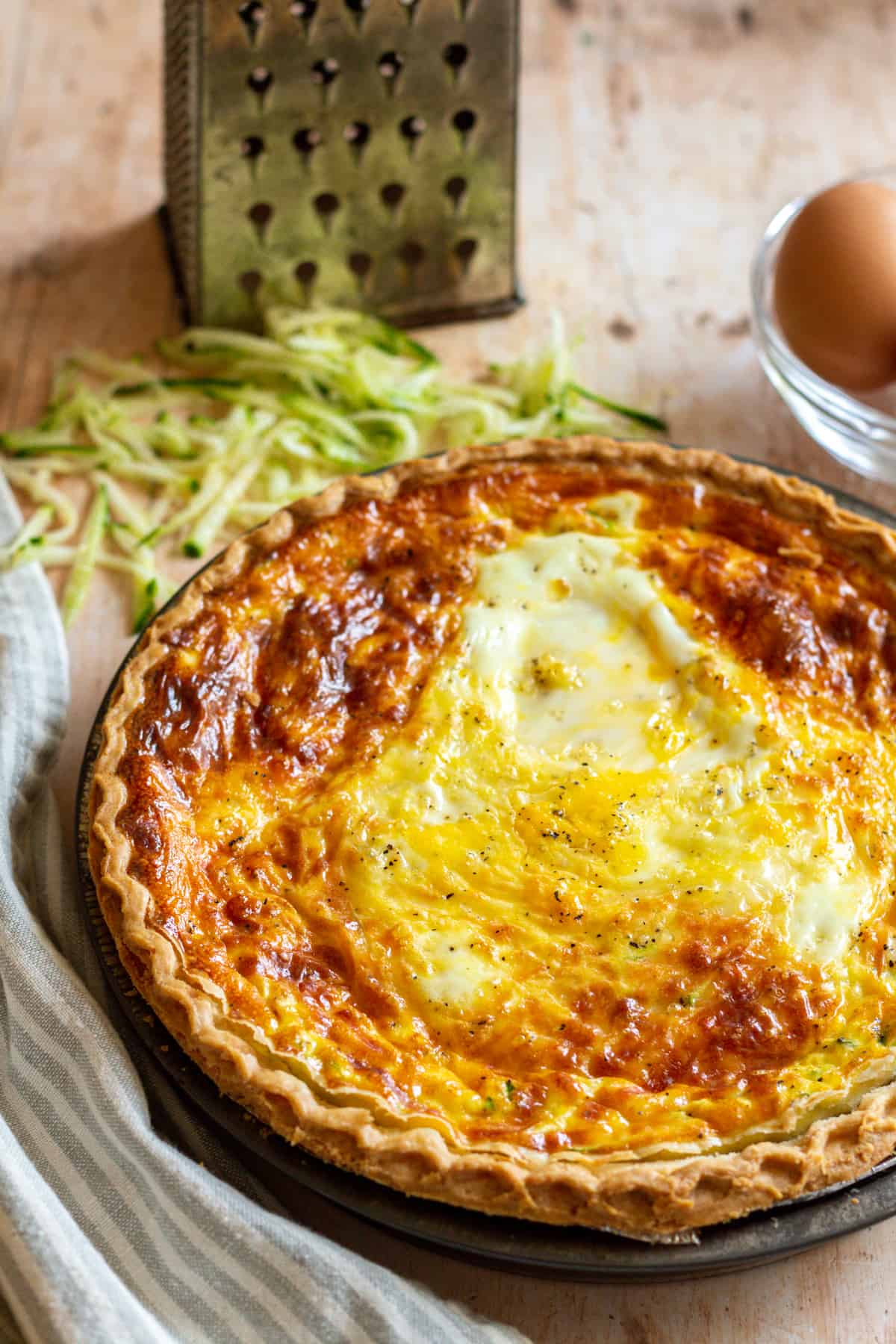 Zucchini quiche in a grey pie tin with a striped napkin, shredded zucchini, and eggs in the background.