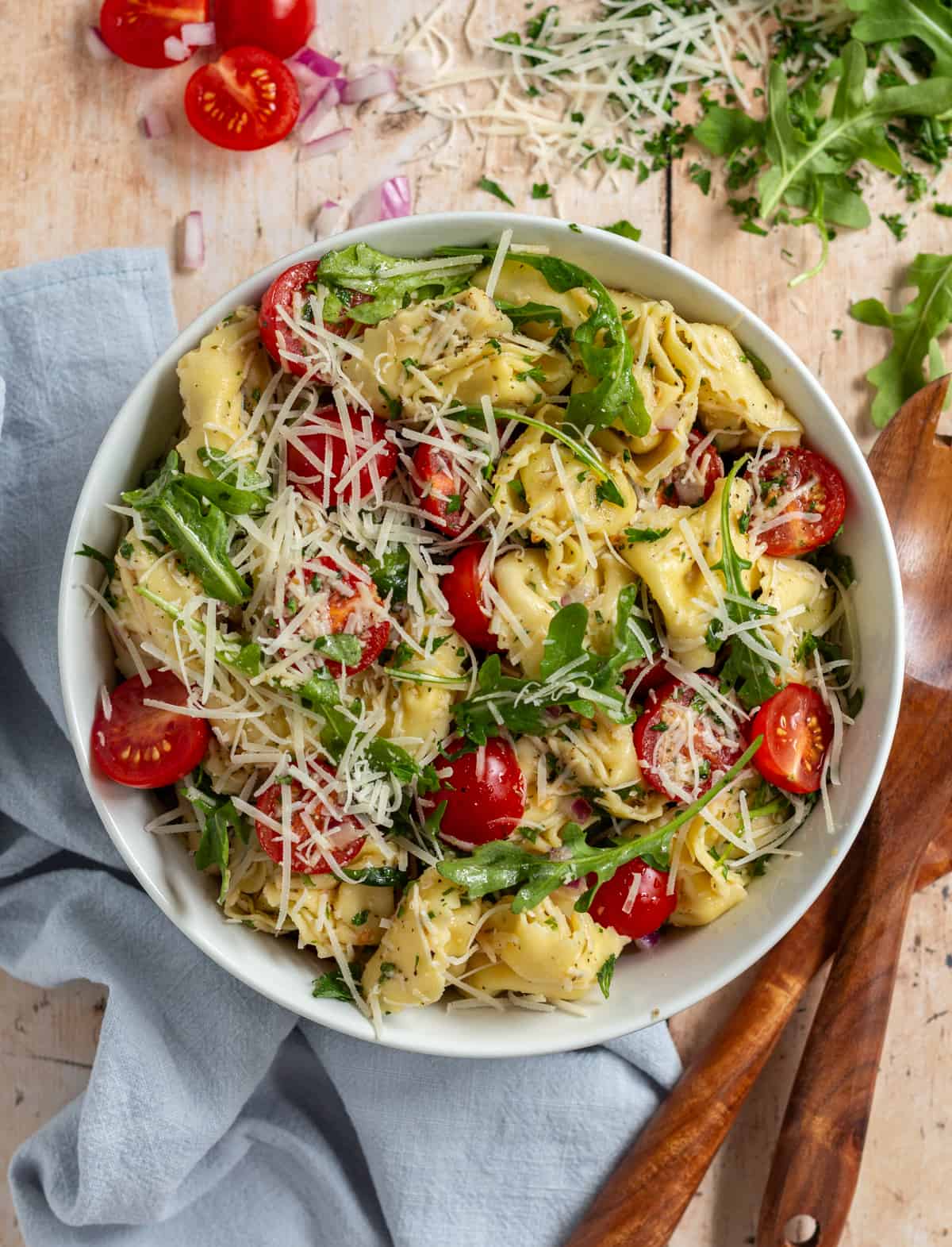 Summer Tortellini Salad topped with cherry tomatoes, arugula, and parmesan cheese. In a white bowl, on a blue napkin, with wooden serving spoons and various ingredients in the background.