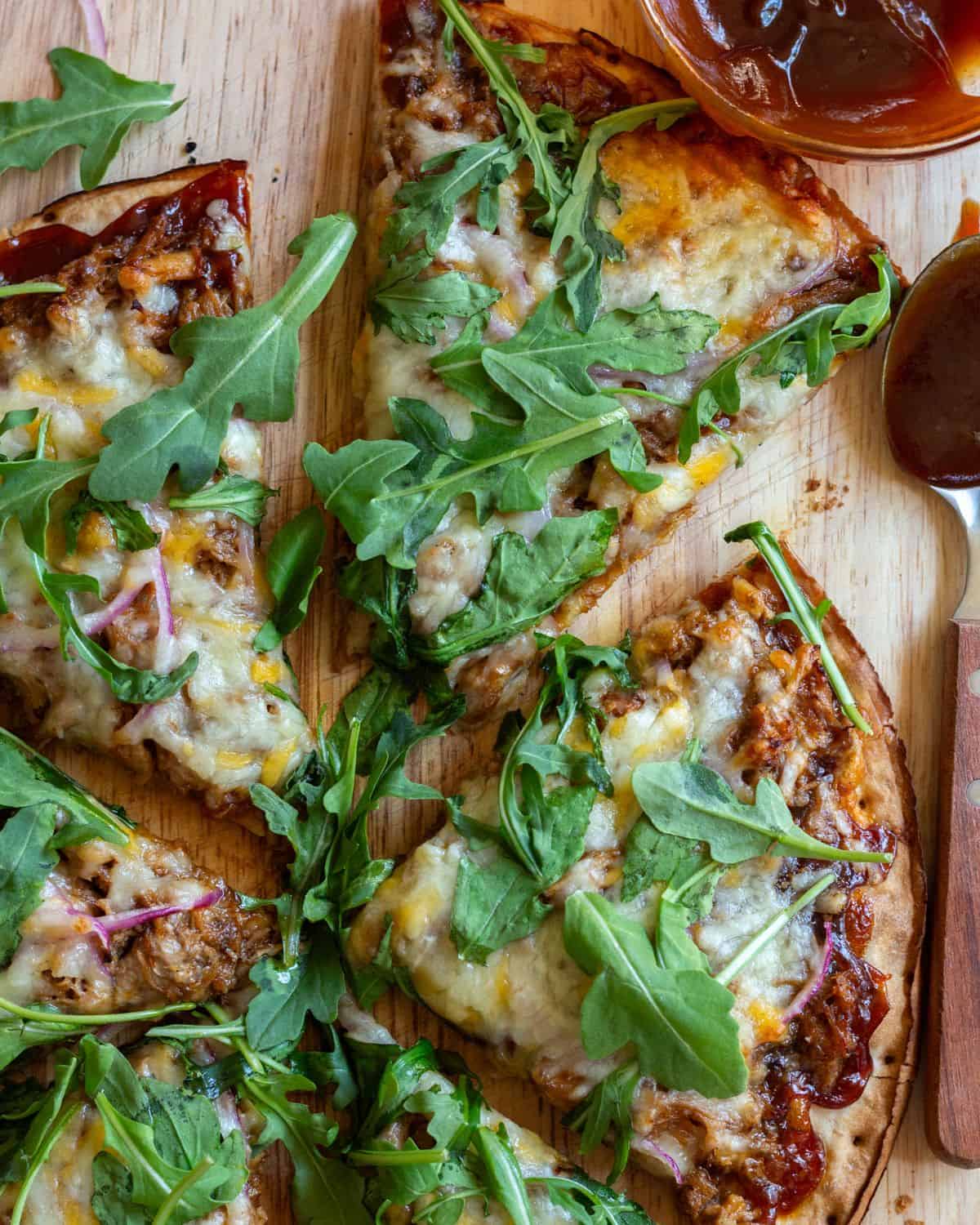 BBQ pulled pork pizza on a cutting board, topped with arugula, with BBQ sauce on the side.