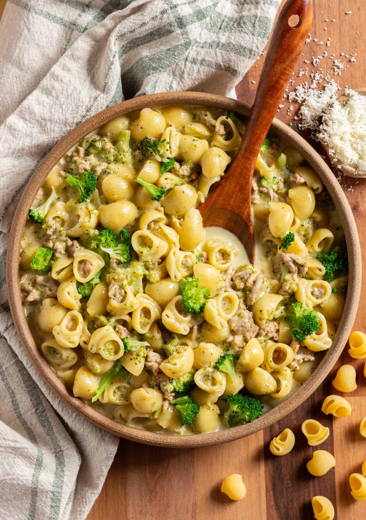 A simple ground turkey pasta recipe with broccoli and cheddar cheese, in a brown bowl with a wooden spoon, sitting on a green and cream colored dish towel.