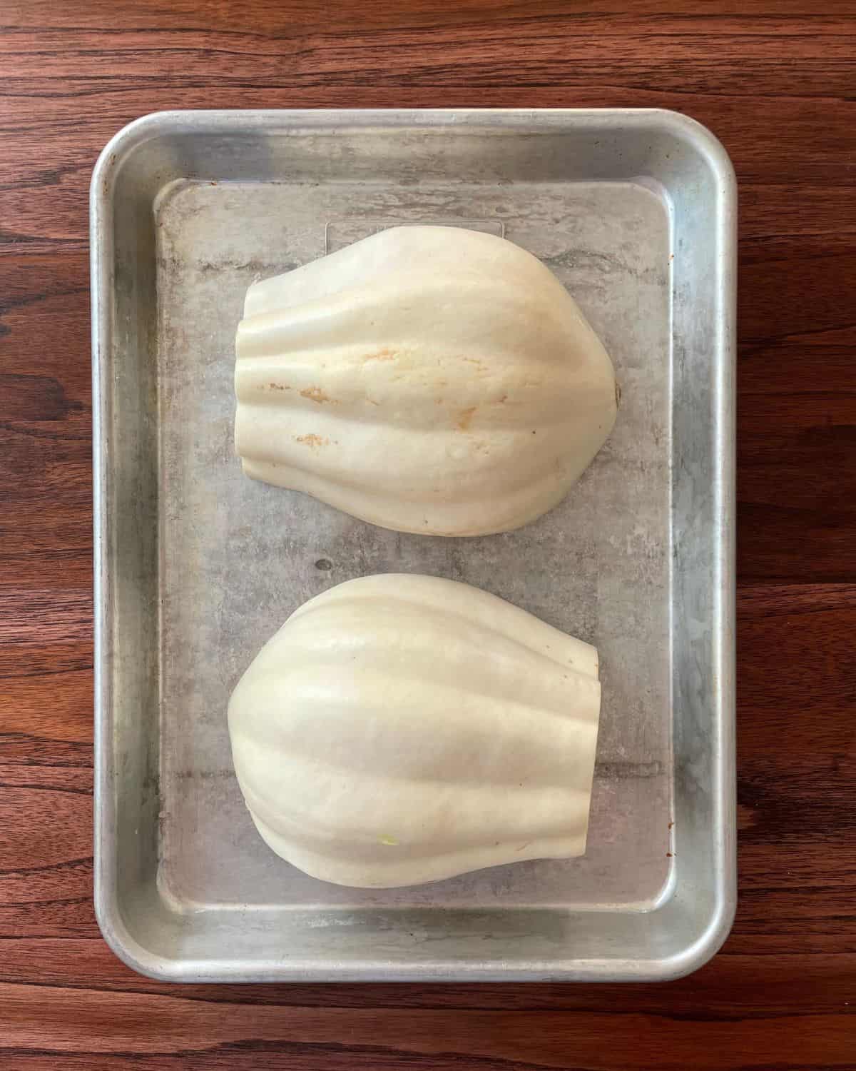 Uncooked white acorn squash cut open with the cut side down, on a baking sheet.