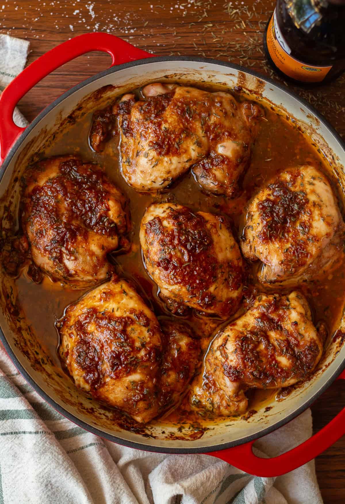 Balsamic chicken thighs, cooked, in a red baking dish with handles, a kitchen towel in the bottom left corner, and jar of balsamic vinegar in the top right corner.