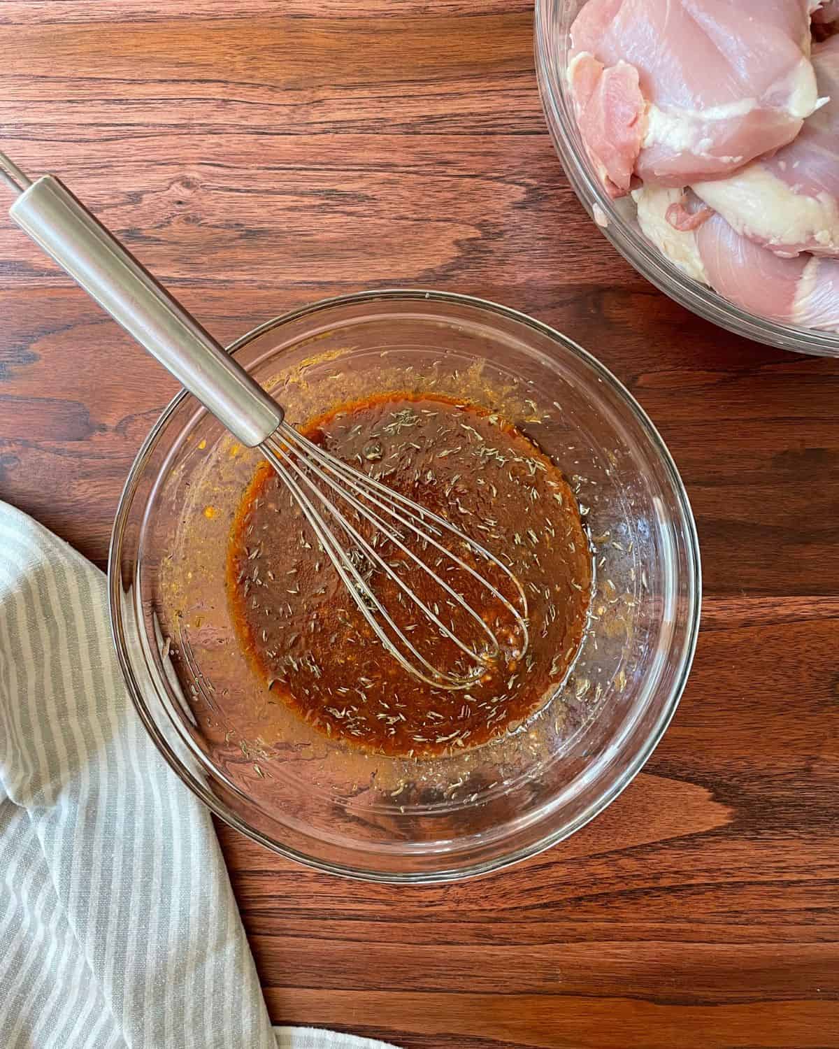 Balsamic marinade whisked together in a glass bowl, with a kitchen towel in the bottom left corner, and a bowl of raw chicken thighs in the top right corner.