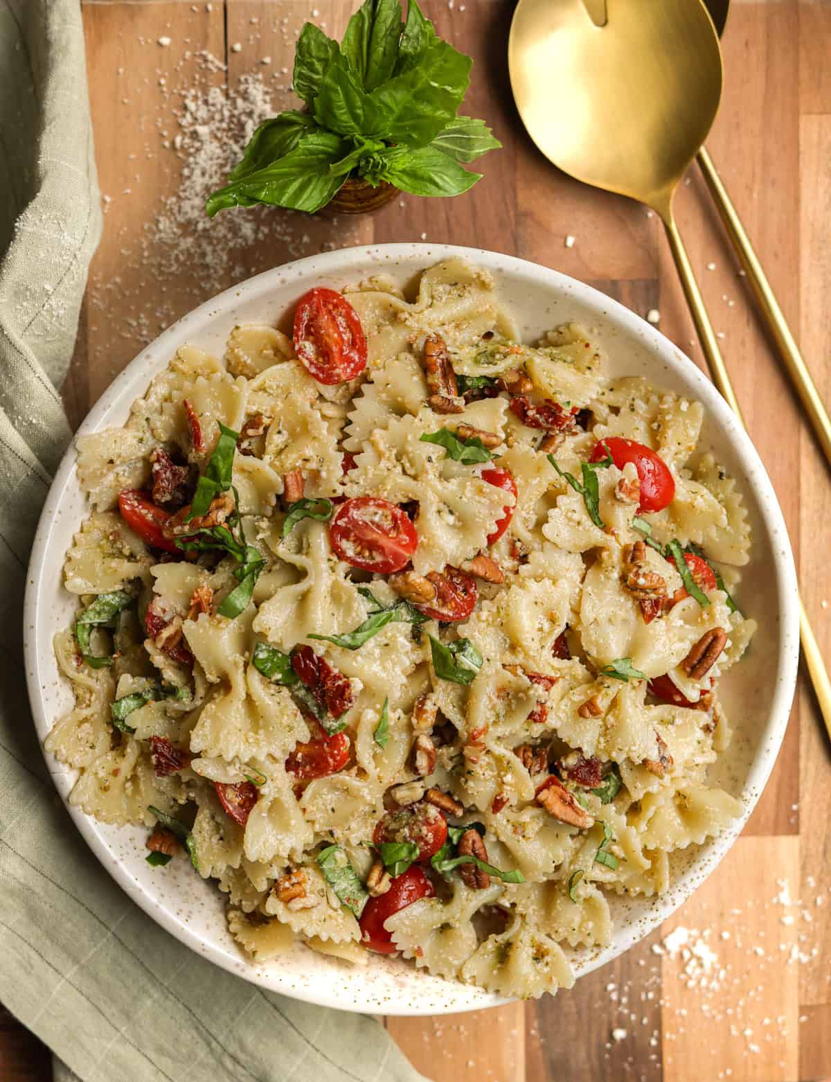 Pesto pasta salad with sundried tomatoes in a serving bowl, on a wooden cutting board, with large gold metal serving spoons.