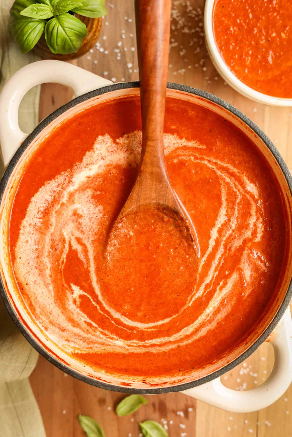 Roasted red pepper and tomato soup (made with canned tomatoes) in a pot with a wooden spoon, garnished with a drizzle of heavy cream.