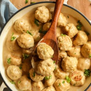 Ground chicken Swedish meatballs in a skillet with a wooden spoon scooping up a couple of meatballs.