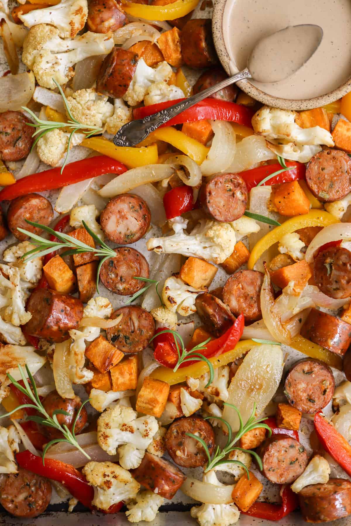 Sheet pan veggies and chicken sausage after it has finished roasting, garnished with fresh rosemary, and a small dish of creamy dressing in the top right corner.