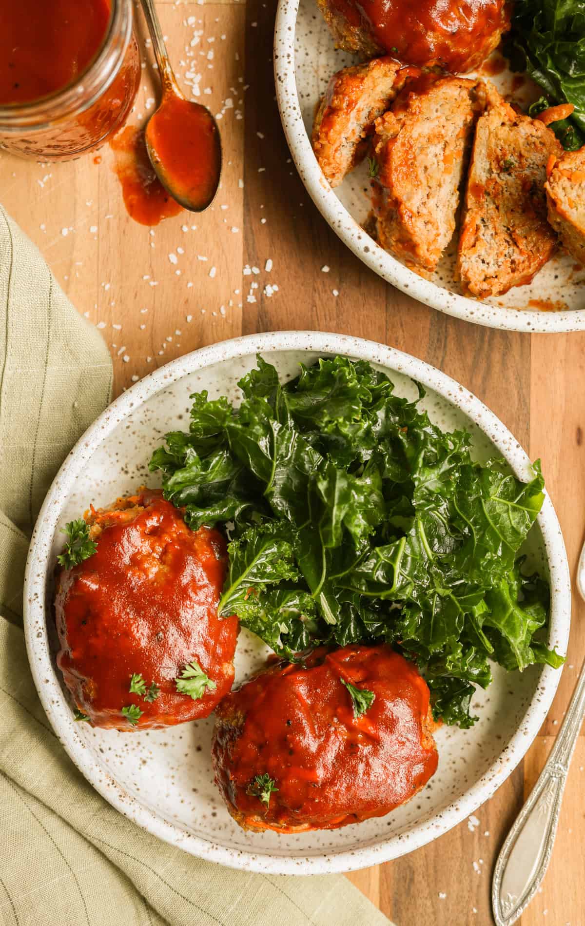 Two ground turkey mini meatloaves on a plate with served with kale. A second plate of sliced meatloaf is in the top right corner and a jar of BBQ sauce is in the top left corner.