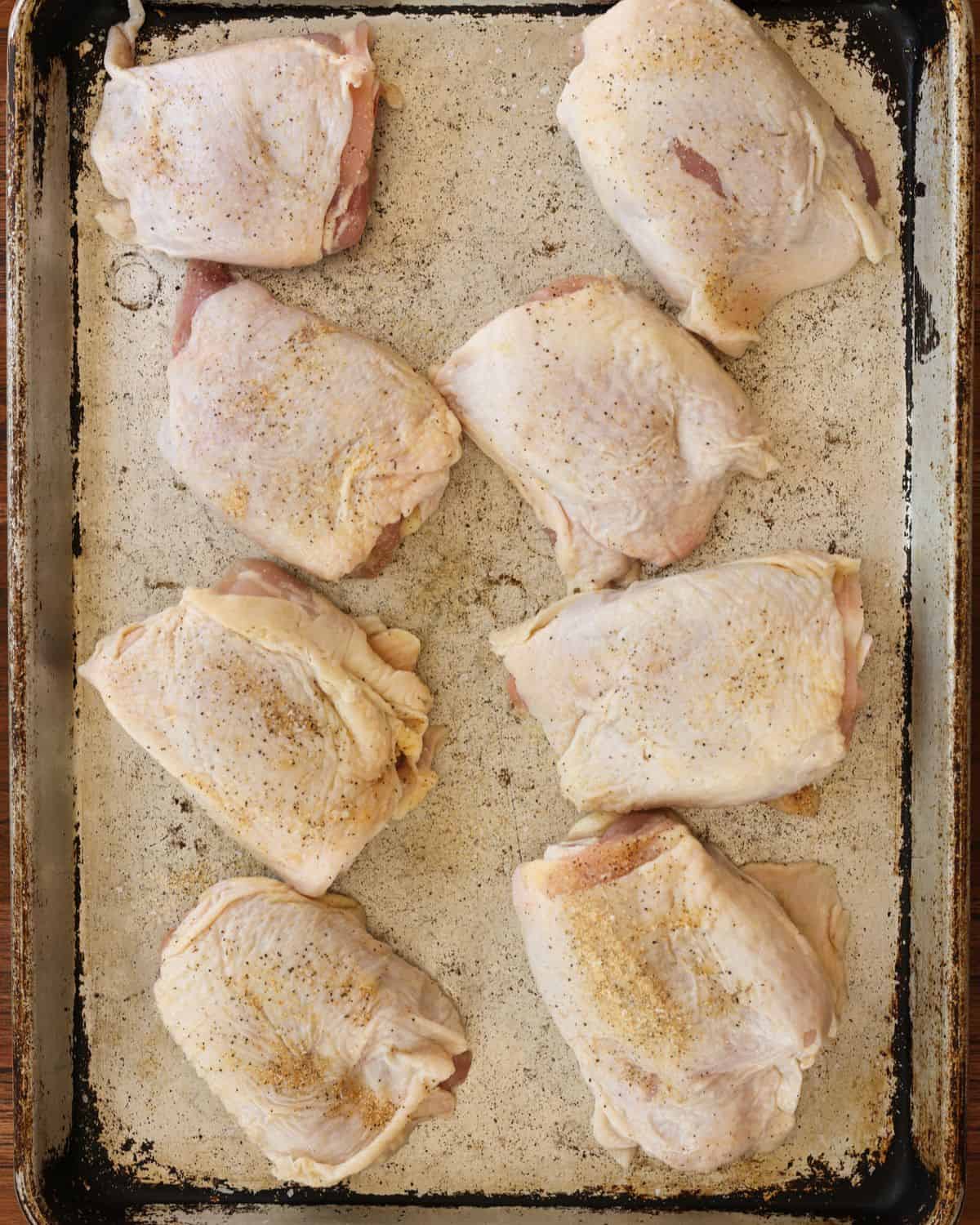 Raw chicken thighs seasoned with salt, pepper, and garlic powder on a baking sheet.